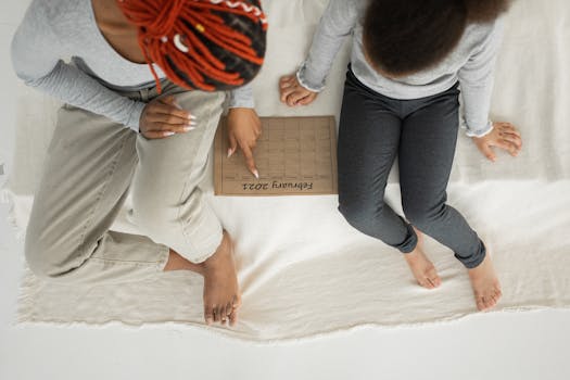 From above of crop anonymous African American female in casual clothes sitting on bed with girl and reading textbook