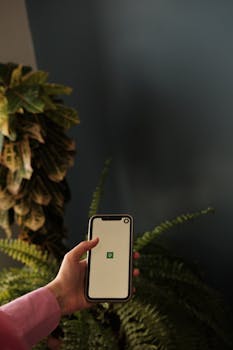A hand holds a smartphone showing an app screen, surrounded by lush indoor plants.